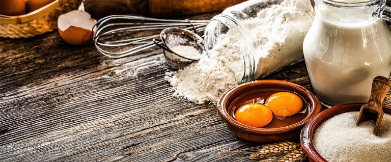Zutaten für ein Backrezept: Mehl, Zucker, Milch, Eier und Küchenutensilien auf einer rustikalen Holzoberfläche.