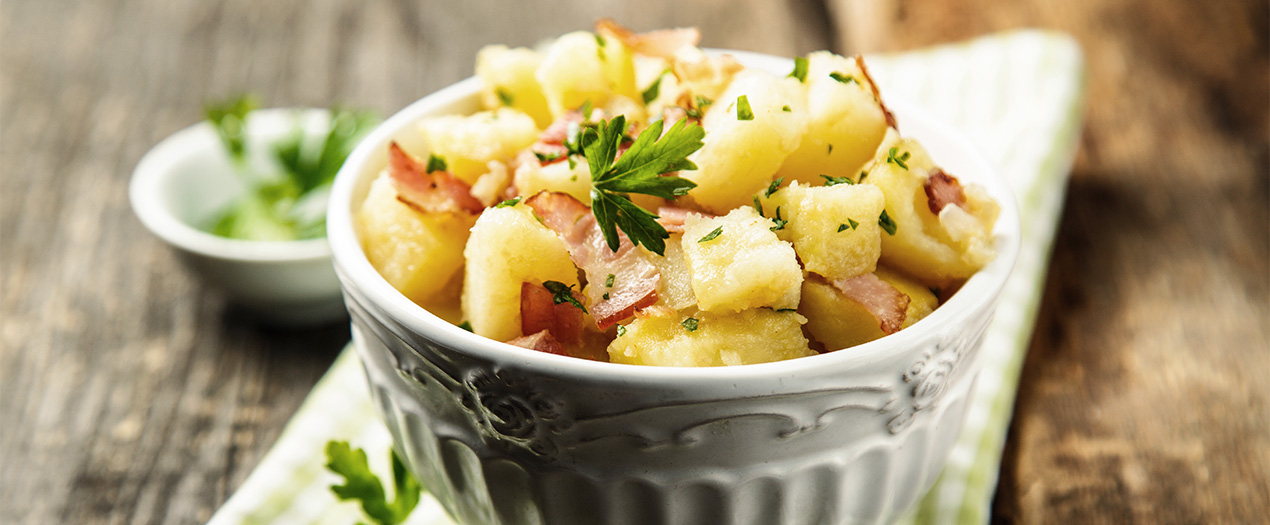 Klassischer Kartoffelsalat mit Speck und frischen Kräutern in einer eleganten Schüssel auf einem rustikalen Tisch.