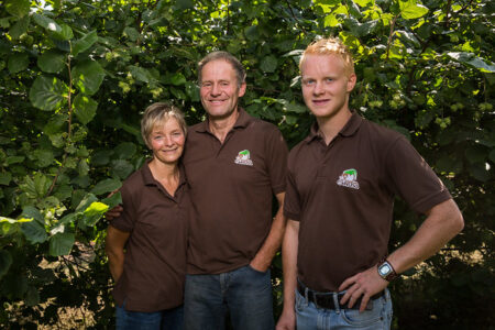 Drei Personen in braunen Shirts stehen lächelnd vor einem Haselnussbaum, symbolisieren familiäre Landwirtschaft.