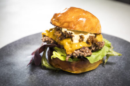 Saftiger Burger mit Wildfleisch, geschmolzenem Käse, Salat und einer glänzenden Brötchenoberfläche auf einem schwarzen Teller.