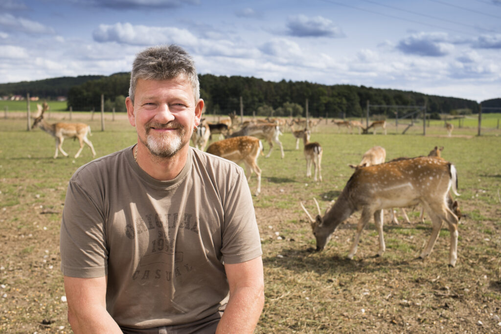 Ein lächelnder Mann sitzt auf einer Wiese mit Rehen im Hintergrund, umgeben von einer idyllischen Landschaft.