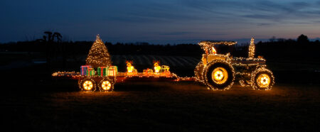 Ein beleuchteter Traktor mit einem Anhänger voller Geschenke und einem Weihnachtsbaum symbolisiert Weihnachten auf dem Land.