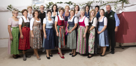Eine Gruppe von Frauen in traditionellen Trachten steht zusammen und lächelt vor einer festlich dekorierten Kulisse.