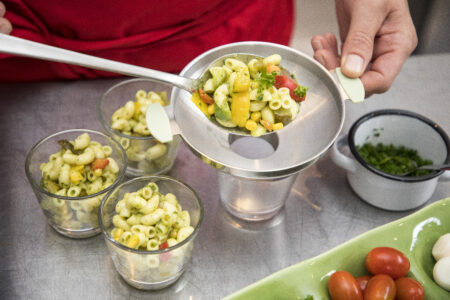 Eine Person füllt mit einem Sieb Pasta-Salat in Gläser, um eine ansprechende Portionierung zu präsentieren.