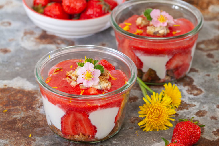 Erdbeerbecher mit frischen Erdbeeren, Joghurt und knusprigem Boden, dekoriert mit Blumen und Kräutern.
