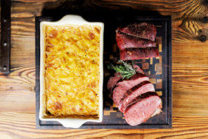 Kartoffelgratin und aufgeschnittenes Fleisch auf einem rustikalen Holzbrett, ideal für Grillgerichte.