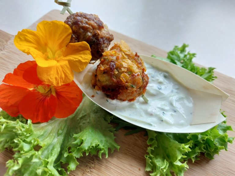 Knusprige Gemüse- und Hackfleischbällchen serviert auf Zaziki-Dip mit frischen Kräutern und essbaren Blumen.