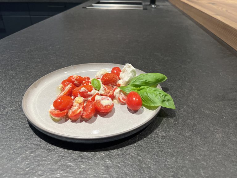Warmer Ziegencamembert mit Kirschtomaten und frischem Basilikum auf einem grauen Teller.
