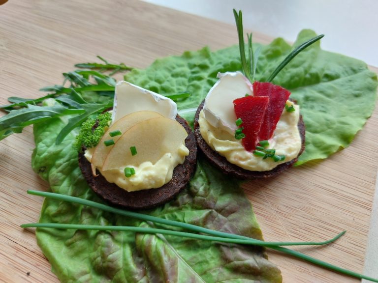 Pumpernickel mit Honig-Senf-Frischkäse, Rote Bete, Apfel und Camembert, garniert mit Kräutern auf einem Salatblatt.