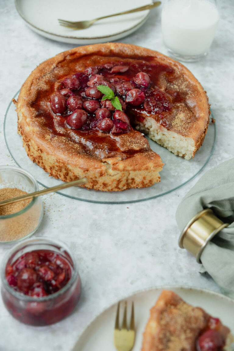 Milchreiskuchen mit Kirschkompott, serviert auf einem Glasuntersetzer mit einer Gabel und Beilagen aus Zucker und Kirschen.
