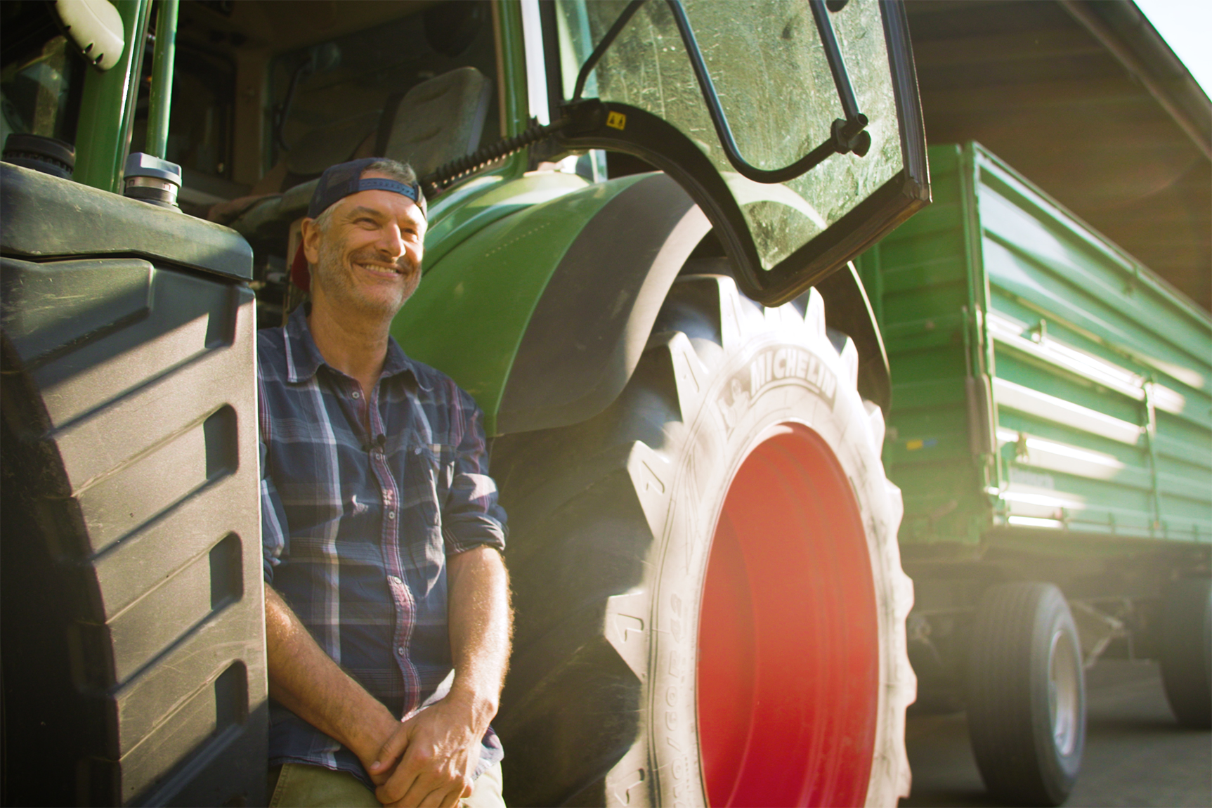 Ein fröhlicher Landwirt lehnt an einem Traktor, während ein grüner Anhänger im Hintergrund zu sehen ist.
