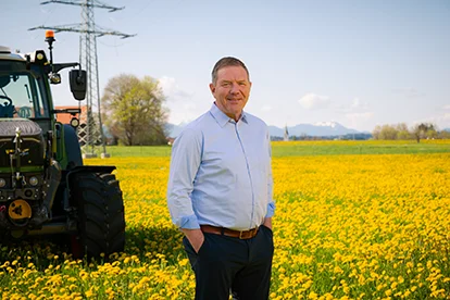 Ein Mann steht lächelnd auf einem blühenden Rapsfeld mit einem Traktor im Hintergrund und Bergen in der Ferne.
