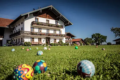 Bunt bemalte Ostereier liegen auf dem grünen Rasen vor einem traditionellen bayerischen Bauernhaus.