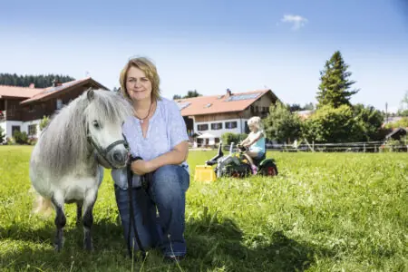 Bäuerin kniet mit Pony auf einer Wiese vor ihrem Bauernhof