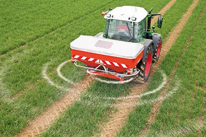 Ein Traktor verteilt Dünger gleichmäßig auf einem grünen Feld mit sichtbaren Spuren im Boden.