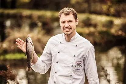 Ein Koch in weißer Uniform hält stolz einen frisch gefangenen Fisch vor einer natürlichen Kulisse.