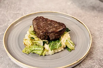 Saftiges Steak auf einem Bett aus cremigem Kartoffelpüree und gebratenem Blattgemüse, serviert auf einem grauen Teller.