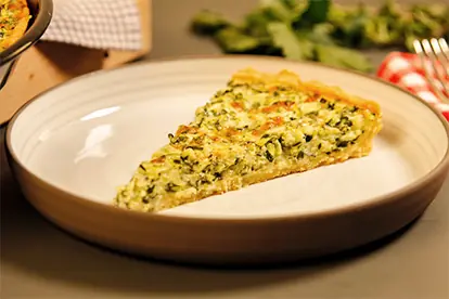 Ein Stück Zucchini-Quiche mit Frischkäse auf einem Teller, umgeben von frischen Kräutern und einem rustikalen Hintergrund.