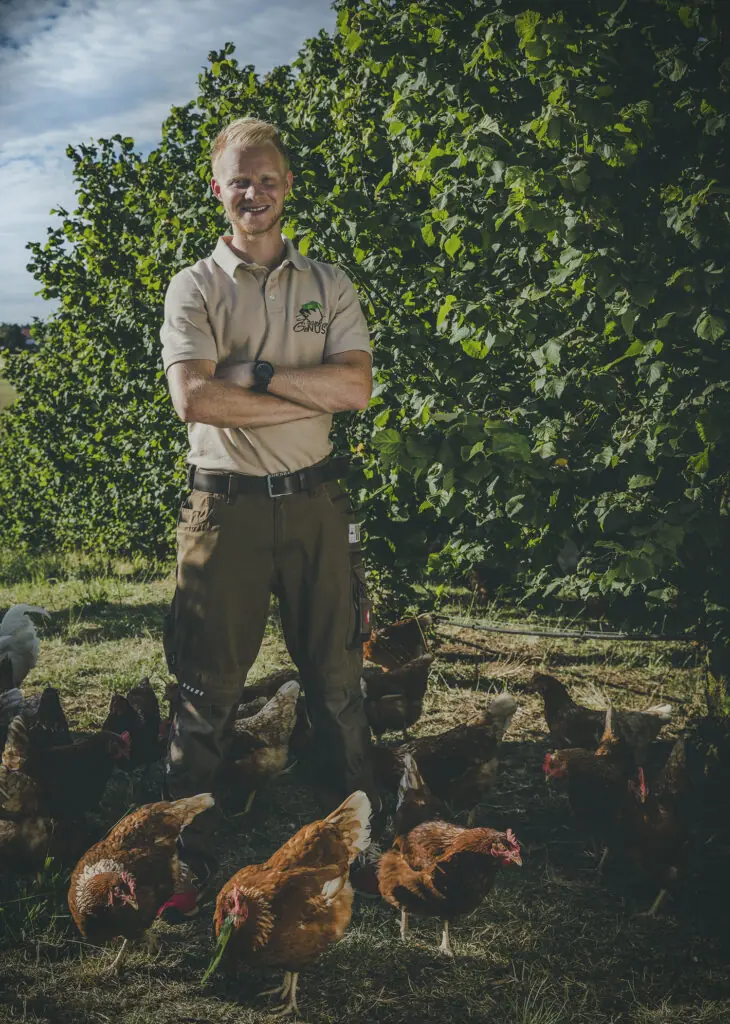 Ein junger Mann lächelt vor einem grünen Hintergrund aus Blättern, trägt ein helles Polo-Shirt mit Logo.