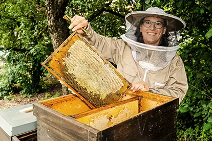 Eine Person in Schutzkleidung zeigt einen Rahmen mit frischem Honig aus einem Bienenstock in einer grünen Umgebung.