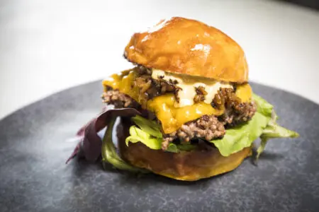Saftiger Burger mit Wildfleisch, geschmolzenem Käse, Salat und einer glänzenden Brötchenoberfläche auf einem schwarzen Teller.
