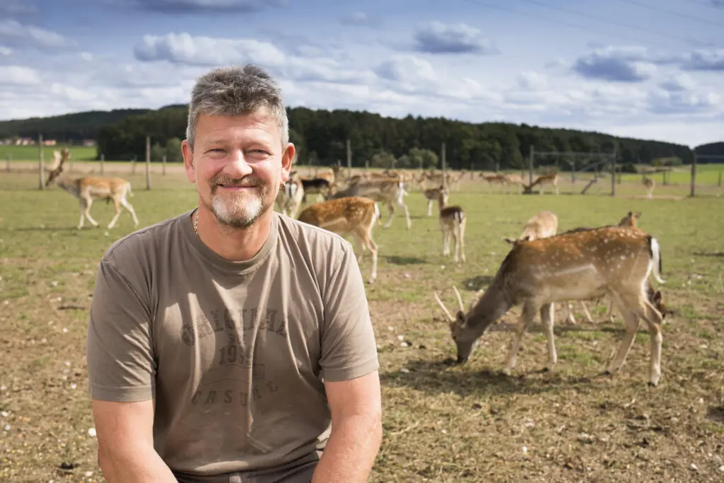 Ein lächelnder Mann sitzt auf einer Wiese mit Rehen im Hintergrund, umgeben von einer idyllischen Landschaft.