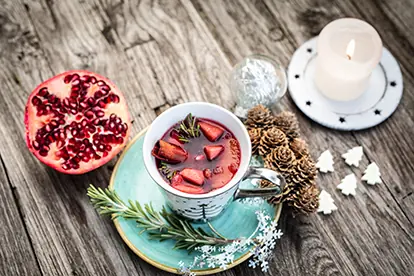 Weihnachtliche Sangria in einer Tasse, umgeben von Granatapfel, Tannenzapfen und einer Kerze auf rustikalem Holz.