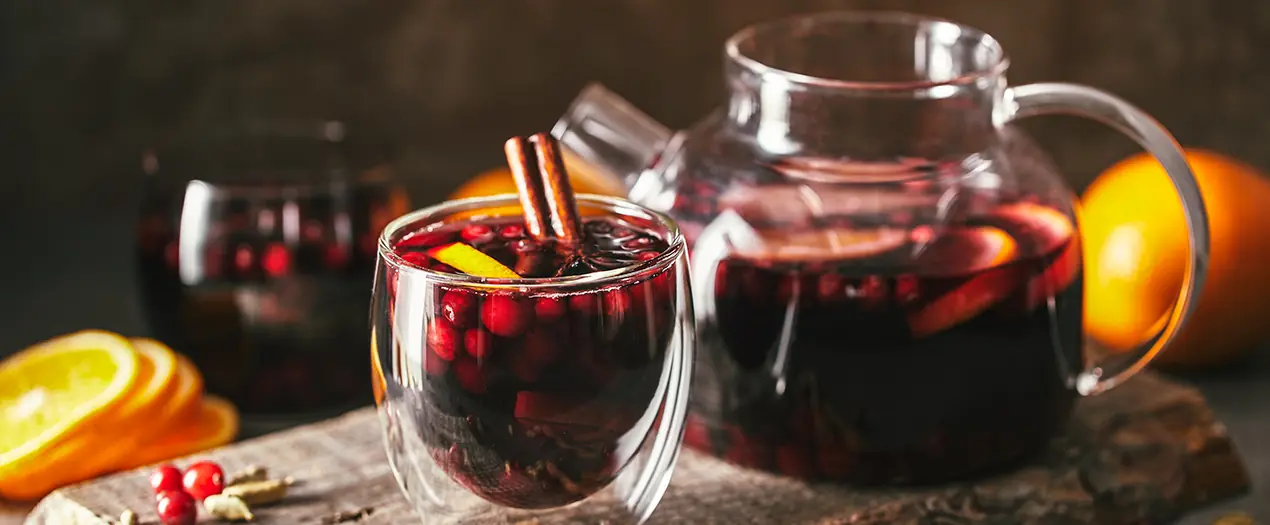 Ein Glas Glühwein mit Zimt und Früchten, daneben eine Kanne und frische Orangen auf einem Holzteller.
