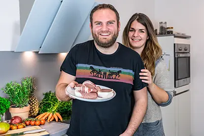 Ein lächelndes Paar hält einen Teller mit Fleisch in einer modernen Küche, umgeben von frischem Gemüse und Zutaten.