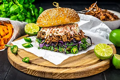 Saftiger Pulled Pork Burger mit Krautsalat, Jalapeños und Limetten auf einem Holzbrett, umgeben von frischen Zutaten.