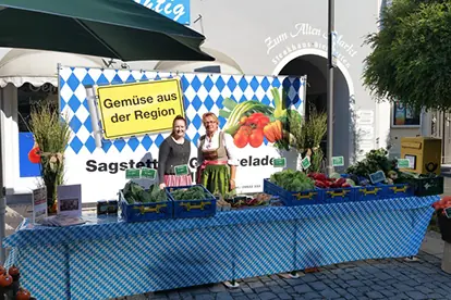 Zwei Verkäuferinnen präsentieren frisches Gemüse aus der Region an einem Marktstand mit bayerischem Dekor.