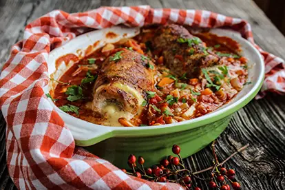 Schweineröllchen in Tomatensauce mit frischen Kräutern, serviert in einer grünen Auflaufform auf rustikalem Holz.