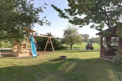 Spielplatz mit Rutsche und Schaukel, umgeben von Bäumen und einem Holzschuppen, im Hintergrund ein Traktor auf dem Feld.