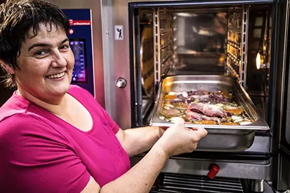 Eine lächelnde Frau holt ein Blech mit Hirschbraten und Gemüse aus dem Ofen.