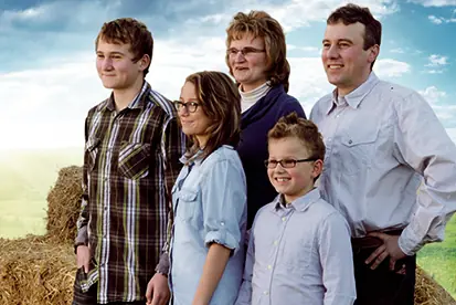 Eine Familie steht lächelnd vor einer ländlichen Kulisse mit Heuballen und blauem Himmel.