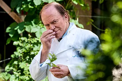 Ein Koch in weißer Uniform steht im Garten und riecht an einem frischen Kräutern, umgeben von grünen Pflanzen.