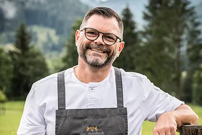 Ein lächelnder Koch in Schürze steht im Freien und präsentiert die traditionelle Zubereitung von Kaspressknödeln.