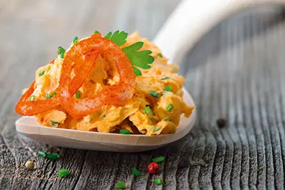 Obatzda auf einem Holzlöffel, garniert mit Paprika und Kräutern, präsentiert sich appetitlich auf rustikalem Hintergrund.