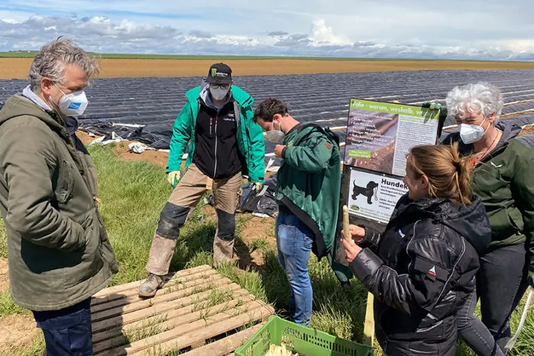 Fünf Personen in Schutzmasken diskutieren auf einem Feld mit einer Informationswand über nachhaltige Landwirtschaft.
