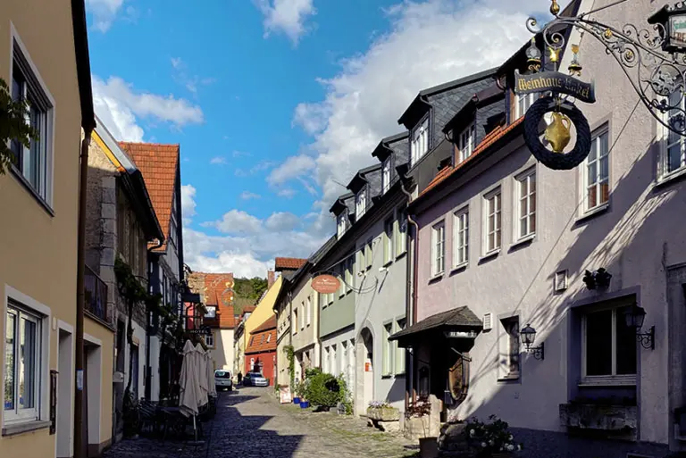 Charmante Gasse mit historischen Gebäuden und einem Weinhaus-Schild, unter blauem Himmel und Wolken.