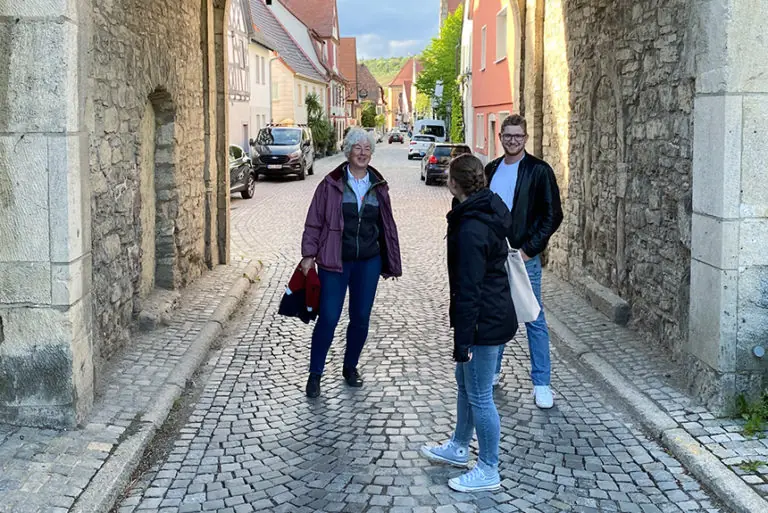 Drei Personen stehen auf einer gepflasterten Straße in einer malerischen Stadt, umgeben von historischen Gebäuden.