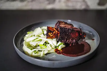 Saftige Rinder-Rippchen serviert mit Spargelsalat und würziger BBQ-Sauce auf einem grauen Teller.
