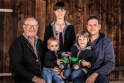 Eine Familie mit Großeltern und Kindern posiert lächelnd vor einer Holzwand, symbolisch für die Heimat der Vielfalt.
