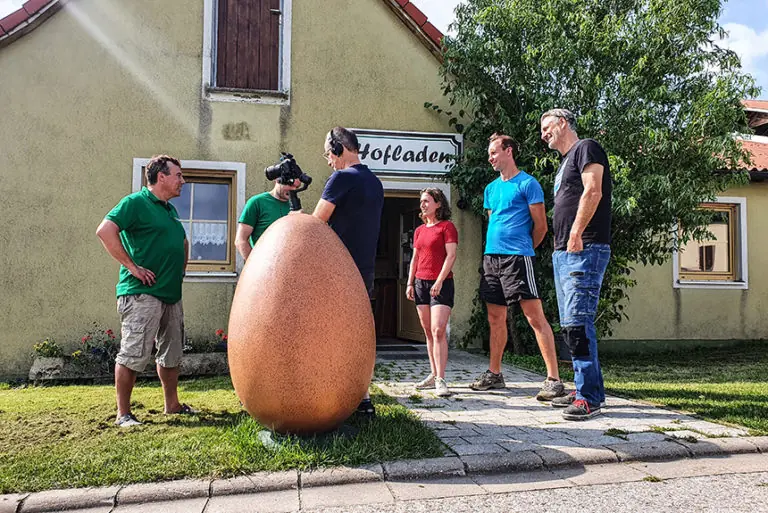 Besucher stehen vor dem Geflügelhof Wellhöfer, um ein großes Ei zu besichtigen und Informationen auszutauschen.