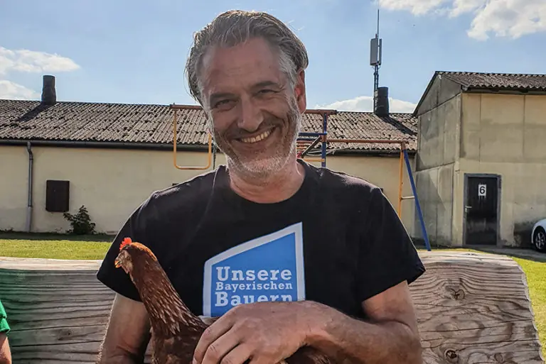 Ein lächelnder Mann mit einem Huhn in der Hand steht auf dem Geflügelhof Wellhöfer, umgeben von ländlicher Kulisse.