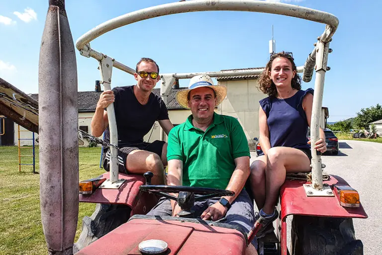 Drei Personen sitzen auf einem Traktor auf dem Geflügelhof Wellhöfer, umgeben von ländlicher Kulisse.