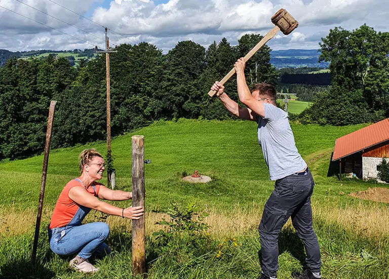 Zwei Personen arbeiten zusammen auf dem Bergbauernhof Zeller, während sie einen Pfosten in den Boden schlagen.