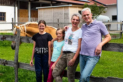 Familie posiert fröhlich vor dem Hof Kuhn mit einem Pferd im Hintergrund, umgeben von grüner Wiese und Holzzaun.