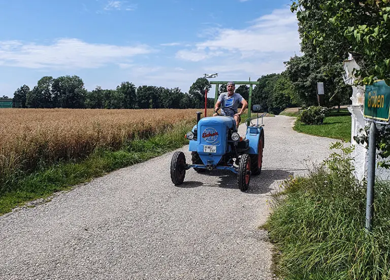 Ein Mann fährt auf einem blauen Traktor auf einem Weg am Steinberger Hof vorbei, umgeben von Feldern und Bäumen.