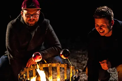 Zwei Männer rösten Äpfel über einem offenen Feuer beim Wintergrillen in einer gemütlichen, nächtlichen Atmosphäre.
